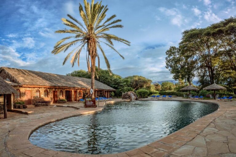 La piscina dell'Amboseli Sopa Lodge, con una palma, ombrelloni di paglia e un edificio in pietra