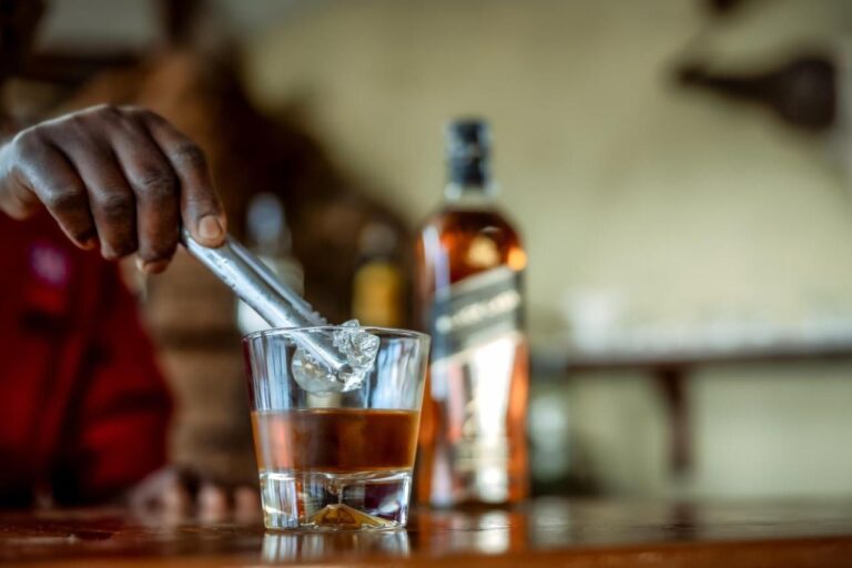 Una mano mette un cubetto di ghiaccio in un bicchiere di whisky, sul bancone del bar
