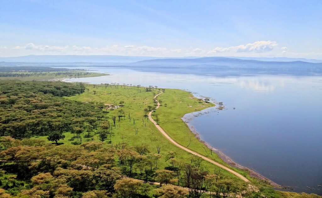 Vista dall'alto del Baboon Cliff sulla riva del lago, una pista che costeggia una prateria verde punteggiata di animali, boschi di acacie e il lago fino alle colline
