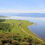 Vista dall'alto del Baboon Cliff sulla riva del lago, una pista che costeggia una prateria verde punteggiata di animali, boschi di acacie e il lago fino alle colline