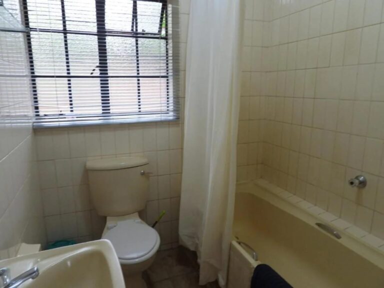 Un bagno piastrellato, con vasca con tenda, WC e lavabo, sotto una finestra con tenda a rullo