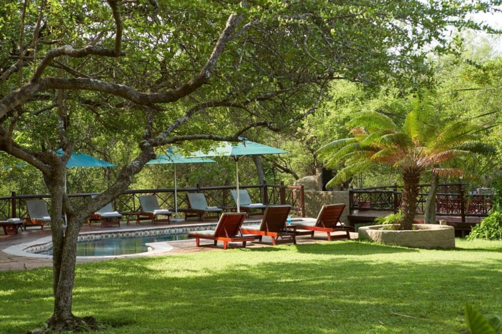 La piscina del Grand Kruger Lodge in un giardino ombreggiato, circondata da lettini in legno e ombrelloni turchesi