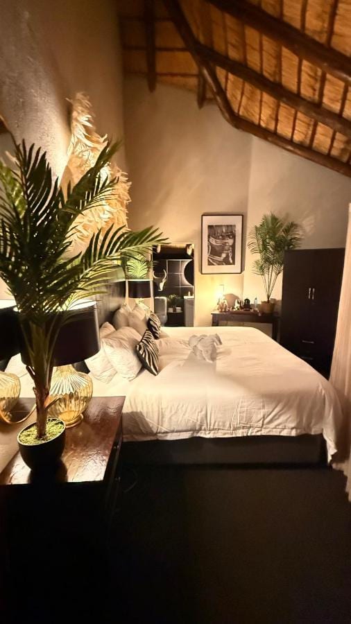 Una camera sotto una travatura di paglia, con un letto matrimoniale bianco, palme in vaso e una foto in bianco e nero alla parete