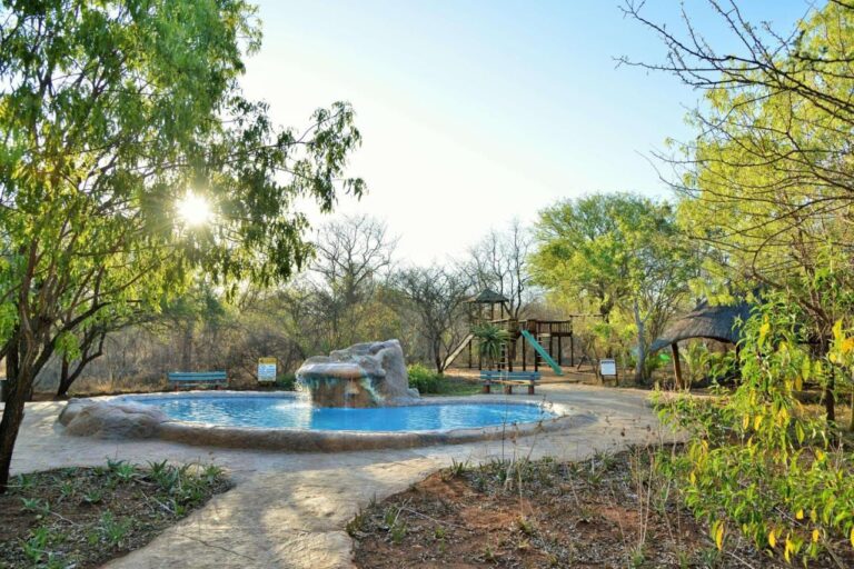 Una piscina con una cascata tra le rocce, davanti al parco giochi e a un riparo di paglia, in controluce
