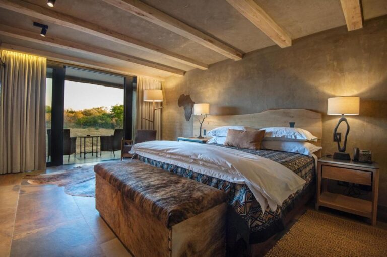 Una suite con un grande letto, una panca in pelle ai piedi del letto e una terrazza arredata davanti alla boscaglia