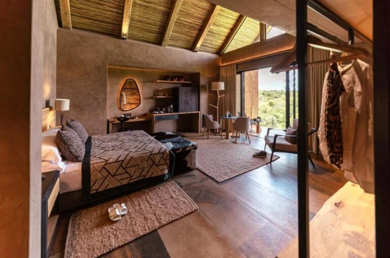 Una suite del Kapama River Lodge sotto una travatura di legno e paglia, con angolo scrivania e vista sugli alberi