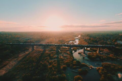 Il treno del Kruger Shalati sul ponte Selati, visto dall'alto, sopra il fiume e la boscaglia, con il sole basso sull'orizzonte