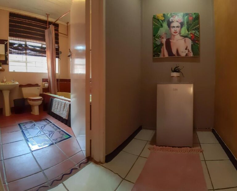 Un bagno con vasca, lavabo e WC, visto da un corridoio dove un piccolo frigorifero sta sotto un ritratto di donna