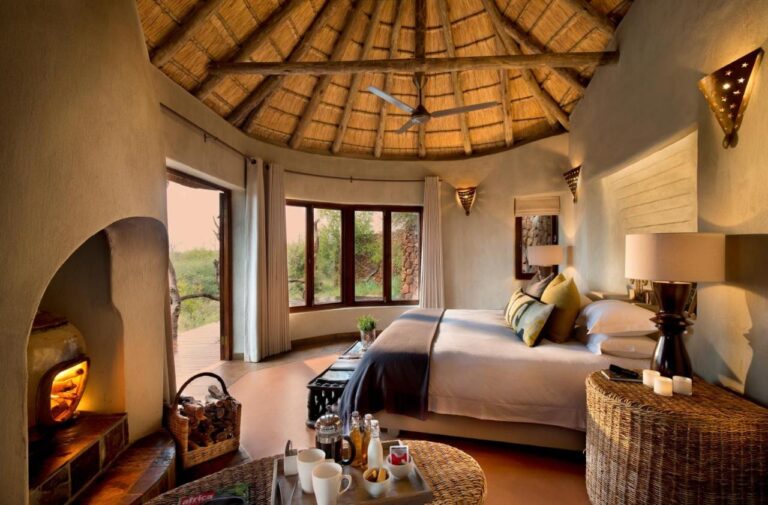Una suite del Madikwe Safari Lodge sotto un tetto di paglia, con un grande letto e un camino acceso