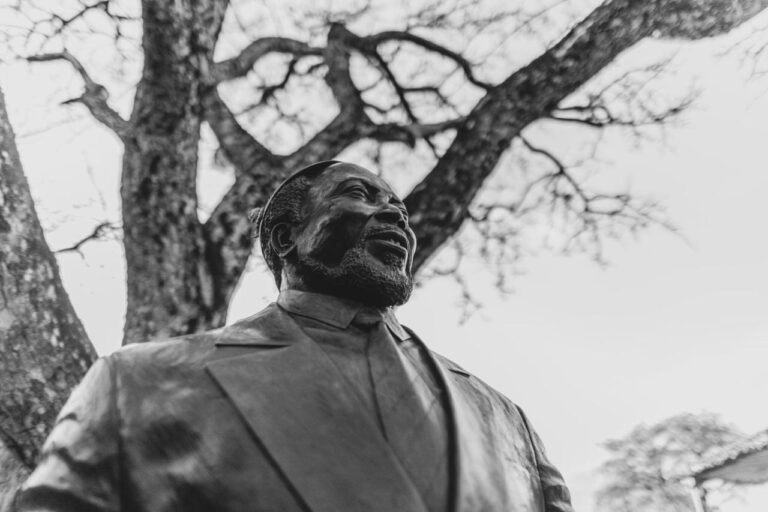 La statua in bronzo di un uomo barbuto e sorridente, in giacca e cravatta, sotto un albero, in bianco e nero