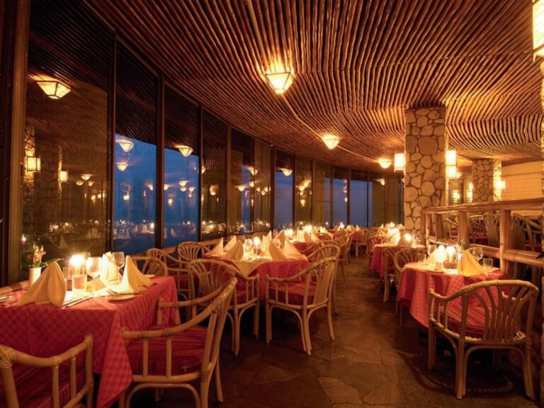 La sala del ristorante a lume di candela, con tovaglie rosse e poltroncine in rattan sotto un soffitto di legno