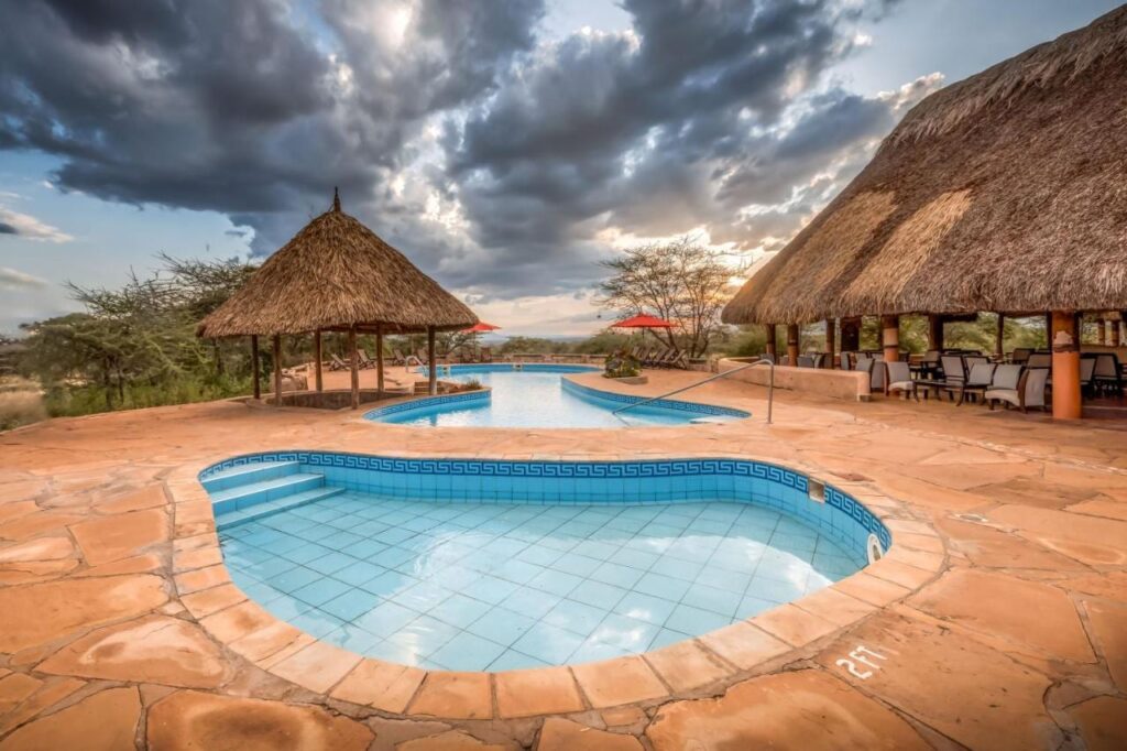 Una piccola vasca piastrellata in primo piano e la grande piscina del Samburu Sopa Lodge, tra un gazebo di paglia e una terrazza con il tetto di paglia