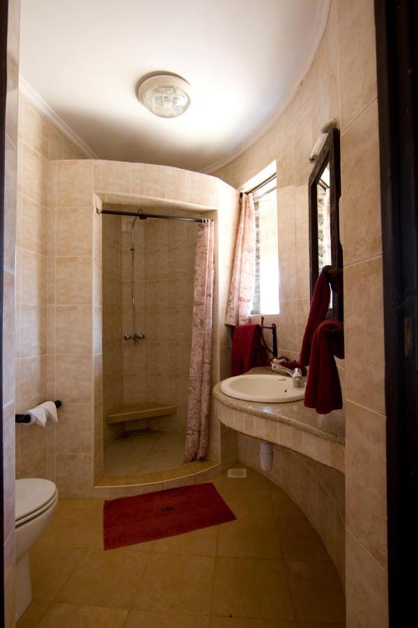 Un bagno piastrellato di beige, con una doccia con tenda, un lavabo su un piano curvo e asciugamani rossi