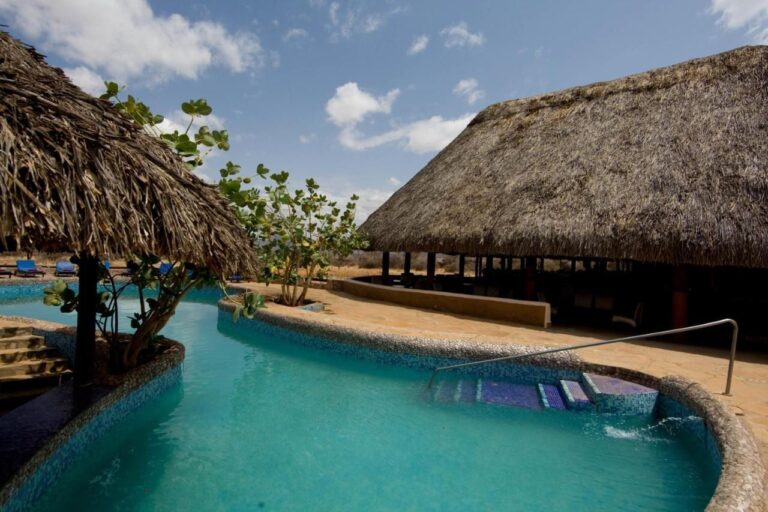 La piscina a mosaico blu, tra un gazebo di paglia e un grande edificio con il tetto di paglia