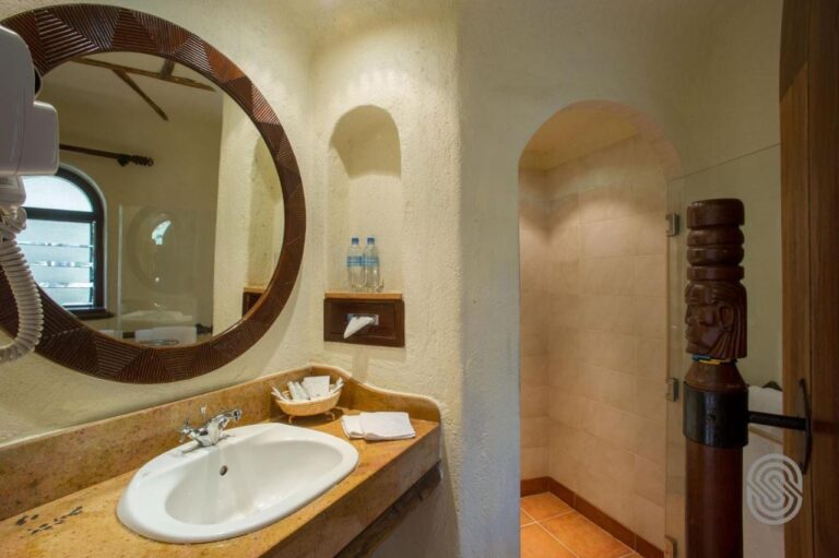 Un bagno con le pareti intonacate, uno specchio rotondo con la cornice in legno intagliato e una doccia sotto un arco