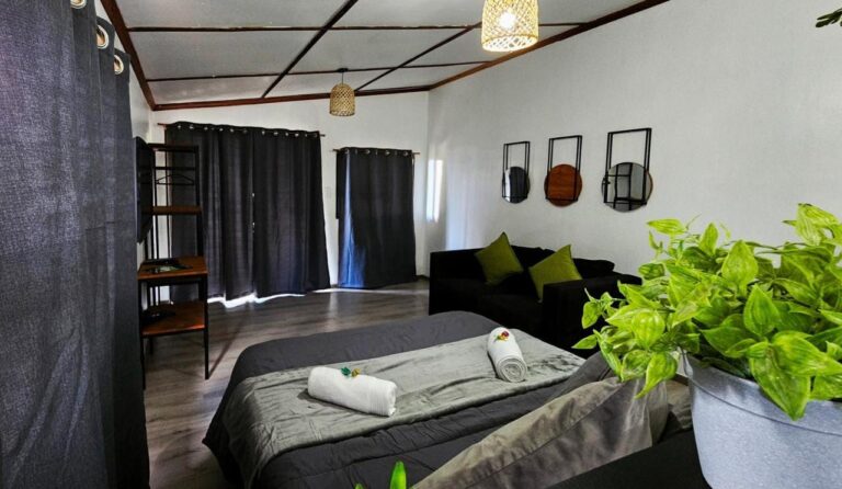 Un letto matrimoniale grigio con asciugamani arrotolati, un divano nero e tende scure, in un monolocale dal soffitto a riquadri di legno