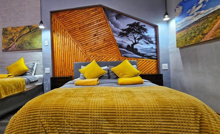 Un letto matrimoniale con plaid giallo davanti a una testiera a listelli di legno e a un dipinto di un albero in bianco e nero, accanto a un secondo letto